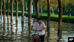 FILE - A man pushes a scooter through floodwaters in Xinxiang in central China's Henan Province, July 26, 2021. 