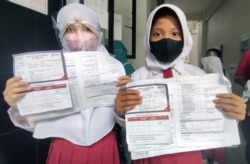 Dua orang siswi menunjukkan kartu registrasi keikutsertaan vaksin untuk anak usia 6 sampai 11 tahun di SD Negeri Cempaka Putih Timur 03. Sebanyak kurang lebih 150 anak berpartisipasi di sekolah dasar tersebut pada 14 Desember 2021. (VOA/Indra Yoga)