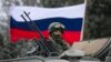 Seorang pria pro-Rusia (tidak terlihat) memegang bendera Rusia di belakang seorang prajurit bersenjata di atas kendaraan tentara Rusia di luar pos penjaga perbatasan Ukraina di kota Balaclava, Krimea, 1 Maret 2014. (Foto: REUTERS/Baz Ratner)